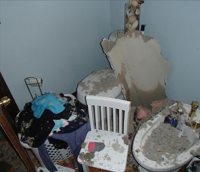 A full laundry hamper, toilet, small chair and sink covered in debris from a caved in ceiling above.