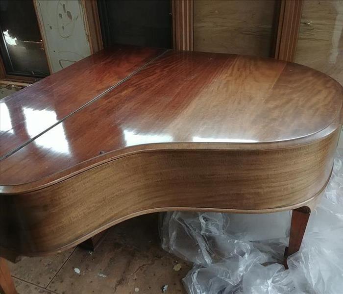 A restored piano with a natural wood finish on top of a plastic sheet in a house that suffered storm damage. 