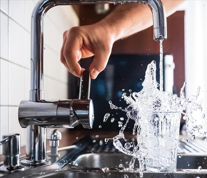 Person pouring overflowing water into glass from kitchen sink.